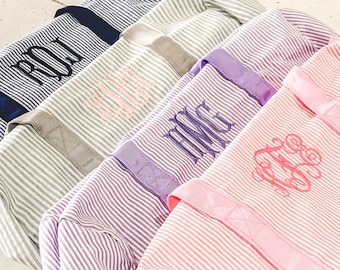 Op de afbeelding: Vier gestreepte canvas tassen met verschillende gekleurde handvatten en gepersonaliseerde monogrammen. De monogrammen zijn geborduurd in verschillende kleuren. De tassen zijn in een rij gerangschikt, met de roze tas vooraan en de blauwe tas achteraan.