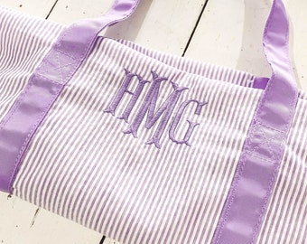Puede incluir: Una bolsa de mano de rayas blancas y moradas con un monograma de las letras "AMG" bordado en la parte delantera.