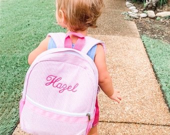 May include: A pink and white striped backpack with the name "Hazel" embroidered on the front. The backpack is being worn by a young child.