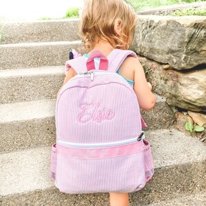 Pode incluir: Uma mochila listrada rosa e branca com fecho de correr e um bolso lateral de malha. A mochila tem o nome "Elsie" bordado na frente.