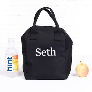 Könnte beinhalten: Schwarze Lunchtasche mit dem Namen "Seth" in weißen Buchstaben. Die Tasche hat einen oberen Griff und eine Seitentasche. Eine Flasche Wasser mit Ananasgeschmack und ein gelb-roter Apfel liegen neben der Tasche.