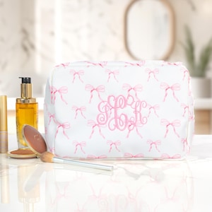 May include: White cosmetic bag with a pink bow pattern and a pink monogram. The bag is rectangular and sits on a white surface with makeup and a brush. The bag is approximately 9 inches long.
