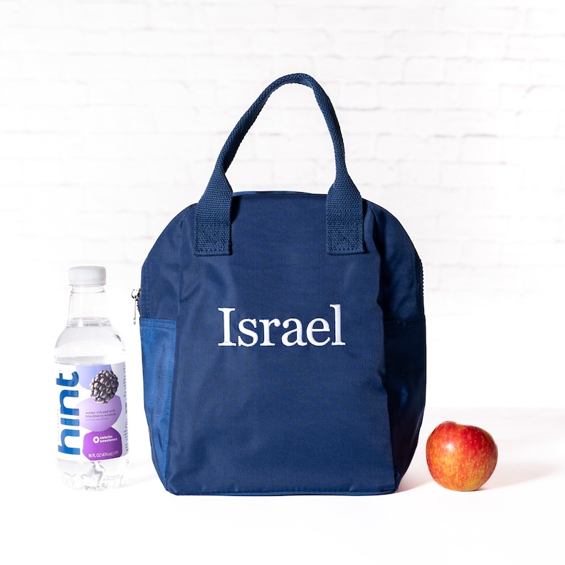 May include: A navy blue lunch bag with a white zipper and the name "Israel" embroidered on the front. The bag is sitting on a white surface next to a bottle of water and a red apple.