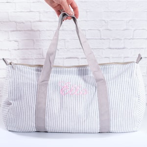May include: A gray and white striped canvas duffel bag with gray handles and a pink embroidered name "Ella".