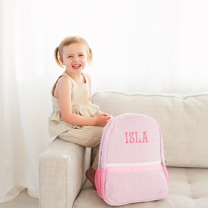 May include: A pink and white striped backpack with a pink zipper and a pink embroidered name "ISLA" on the front. A young girl is sitting on a couch behind the backpack.