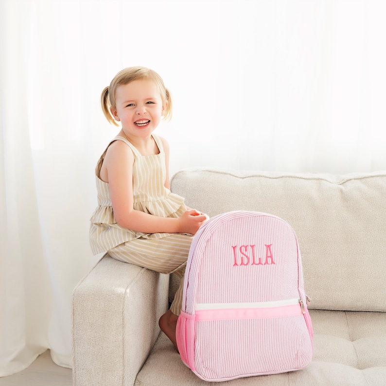 May include: A pink and white striped backpack with a pink zipper and a pink embroidered name "ISLA" on the front. A young girl is sitting on a couch behind the backpack.