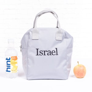 May include: A gray lunch bag with a white zipper and white handles. The bag has the name "Israel" embroidered on the front in black.