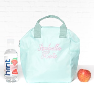 May include: A light blue lunch bag with a zipper closure and a top handle. The bag is personalized with the name "Isabella Kate" embroidered in pink.