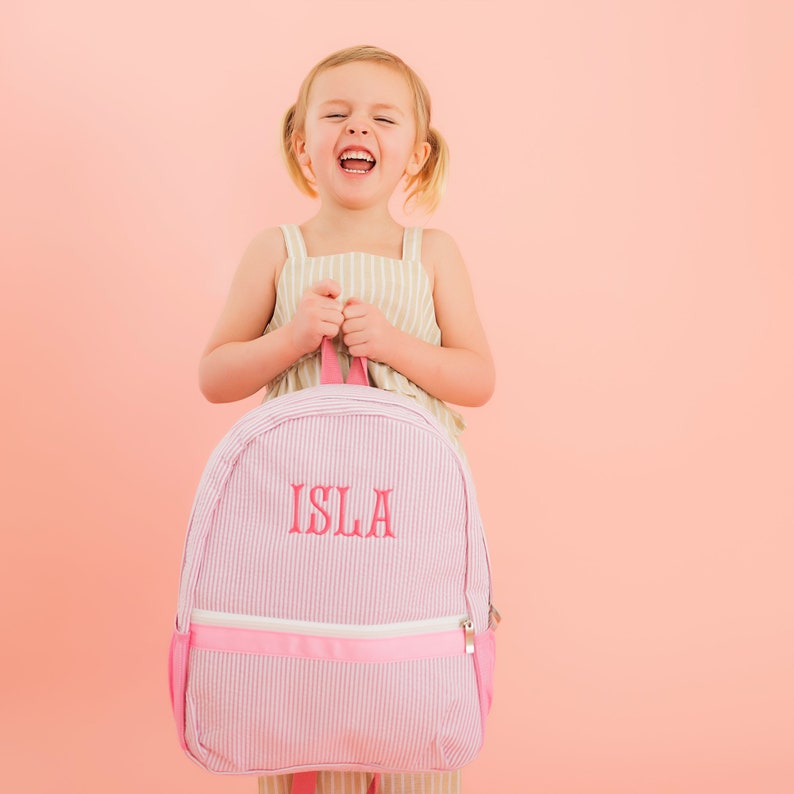 May include: A pink and white striped backpack with a pink zipper and the name "ISLA" embroidered in pink on the front.