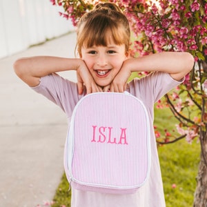 May include: A pink and white striped lunch bag with a white zipper and a pink embroidered name "ISLA".
