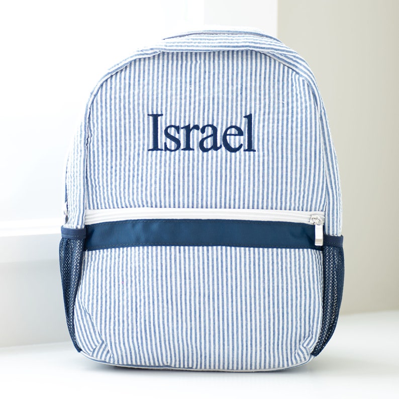 May include: Blue and white striped backpack with a navy blue trim and a zipper closure. The backpack is personalized with the name "Israel" in navy blue embroidery.