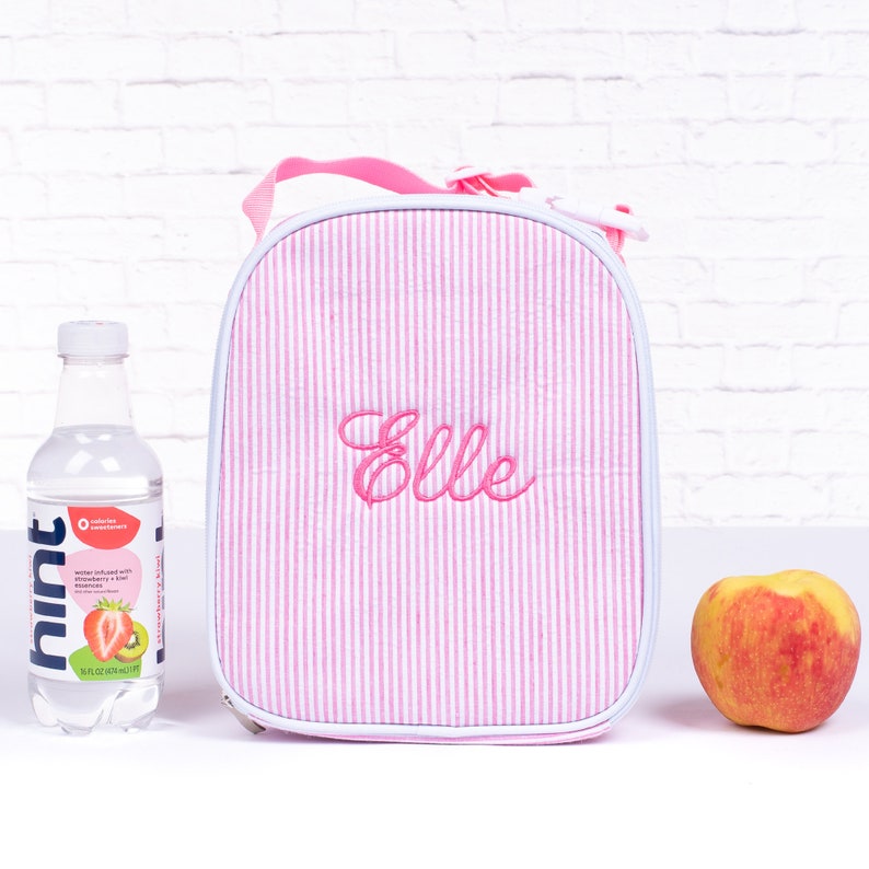 May include: Pink and white striped lunch bag with a white zipper and a pink handle. The bag is personalized with the name "Elle" embroidered in pink.