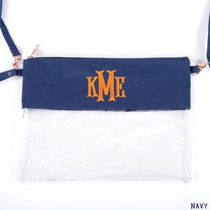 May include: A navy blue clear crossbody bag with a zipper closure and a gold chain strap. The bag has a clear plastic front and a navy blue fabric back with the monogram "KME" embroidered in orange.