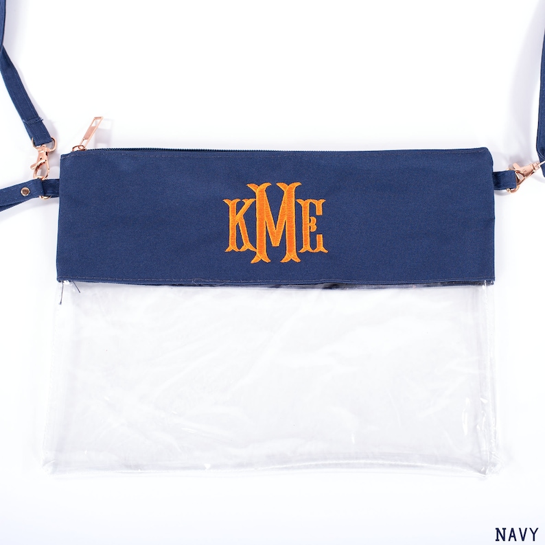 May include: A navy blue clear crossbody bag with a zipper closure and a gold chain strap. The bag has a clear plastic front and a navy blue fabric back with the monogram "KME" embroidered in orange.
