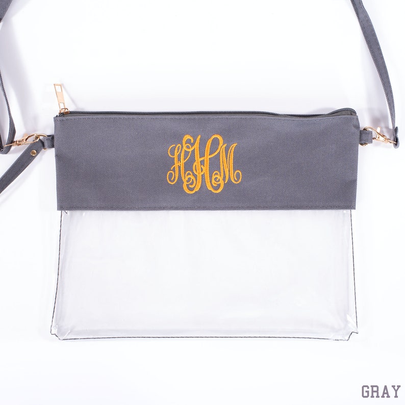 Op de afbeelding: Grijze crossbodytas met een doorzichtige voorkant en een grijs bovenstuk. De bovenkant heeft een gouden rits en een goud geborduurd monogram. De tas heeft een lange, verstelbare grijze riem en goudkleurige hardware. Het woord "GRAY" staat onderaan.