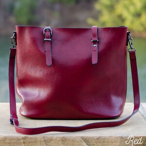 Pu&ograve; includere: Una borsa tote in pelle rosso scuro con tracolla. La borsa ha due manici corti con fibbie argentate e una lunga tracolla regolabile. La parola "Red" &egrave; scritta in bianco in basso a destra.