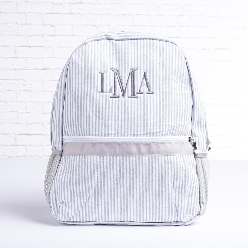 May include: A gray and white striped backpack with a gray zipper and a gray embroidered monogram "LMA" on the front.