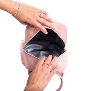 May include: A light pink backpack with a zippered opening. The interior is lined with a silver material. The backpack has a top handle and adjustable shoulder straps. The bag is being opened by a person.