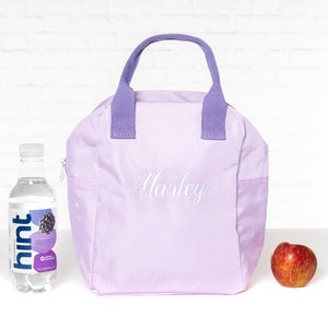 May include: A lavender lunch bag with a purple handle and the name "Marley" embroidered on the front. The bag is sitting on a white surface next to a bottle of water and an apple.