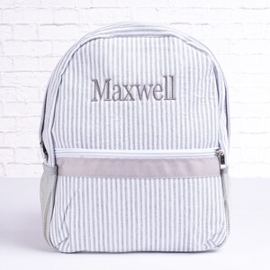 May include: Gray and white striped backpack with a gray zipper and a gray embroidered name "Maxwell" on the front.