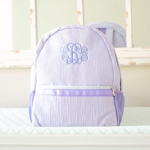 May include: A lavender and white striped backpack with a monogrammed initial "B" in a light blue color.