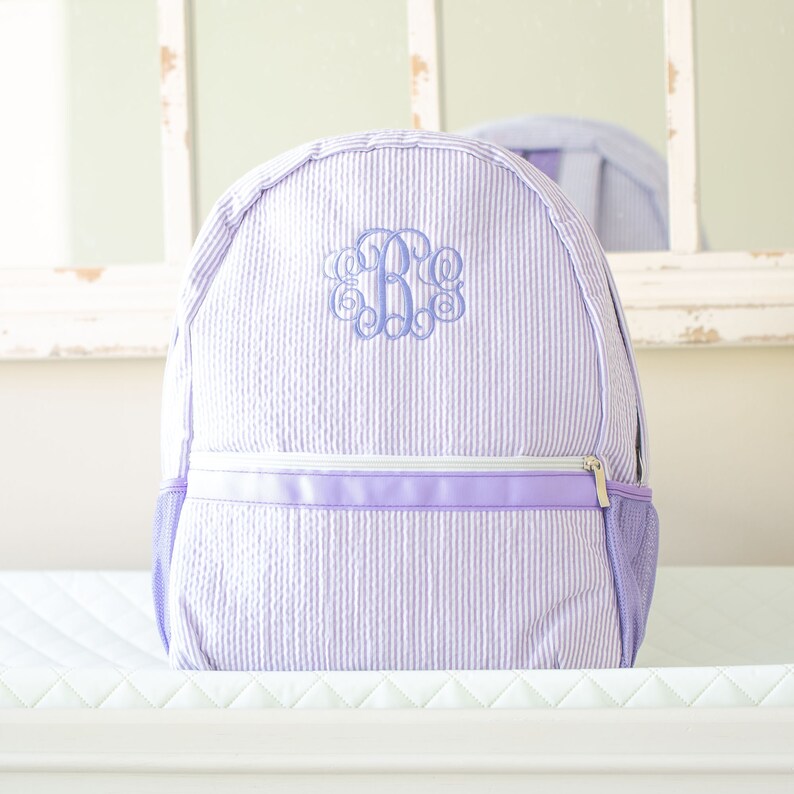 May include: A lavender and white striped backpack with a monogrammed initial "B" in a light blue color.