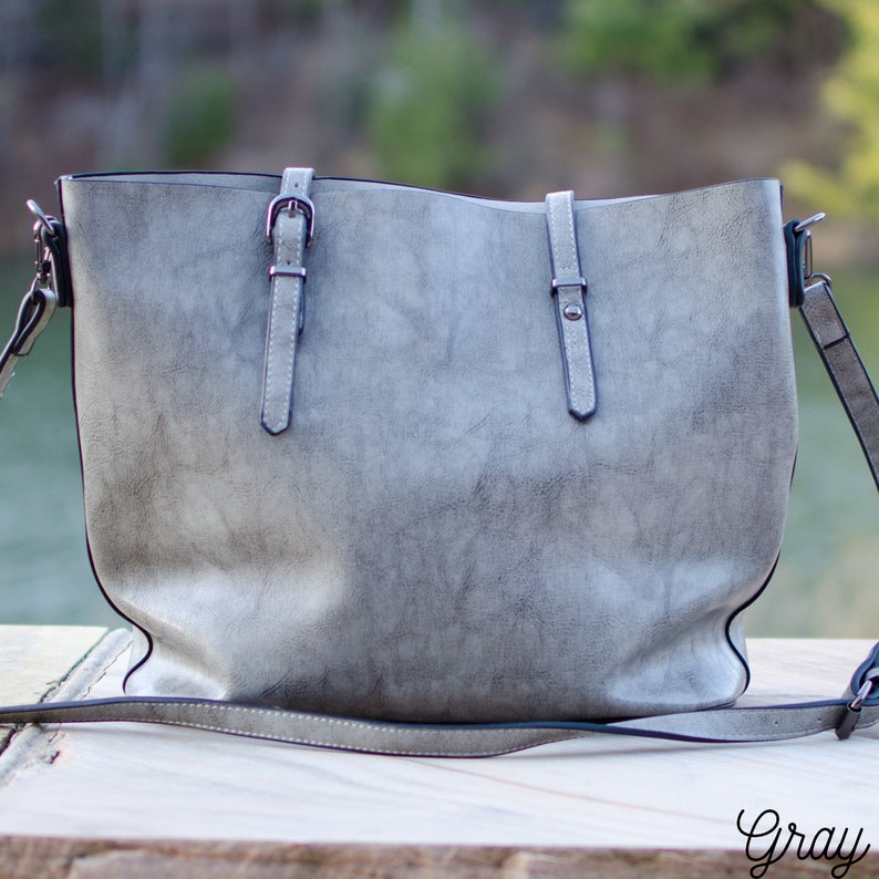 Pu&ograve; includere: Una borsa tote grigia con finitura strutturata. La borsa ha due manici superiori e una lunga tracolla regolabile. Presenta due cinturini decorativi con fibbie sul davanti. La parola "Gray" &egrave; scritta in corsivo in basso a destra.