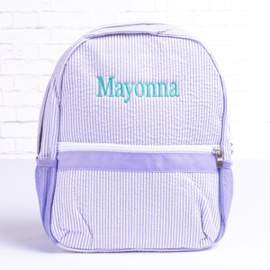 May include: A purple and white striped backpack with a zippered pocket and mesh side pockets. The backpack is personalized with the name "Mayonna" in teal green embroidery.