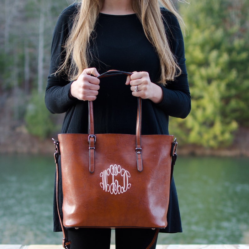 Pu&ograve; includere: Una borsa tote in pelle marrone con monogramma bianco. La borsa ha due manici superiori e una tracolla staccabile. La borsa &egrave; tenuta da una persona che indossa una maglia nera a maniche lunghe.