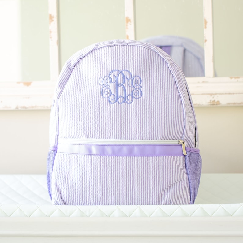 May include: A purple and white striped backpack with a monogrammed initial "B" in the center. The backpack has a zippered pocket and a mesh side pocket.