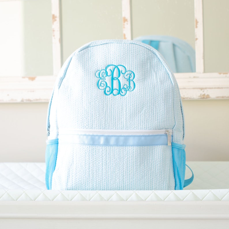 May include: A light blue backpack with a white zipper and a blue monogram on the front. The backpack has a mesh pocket on the side.