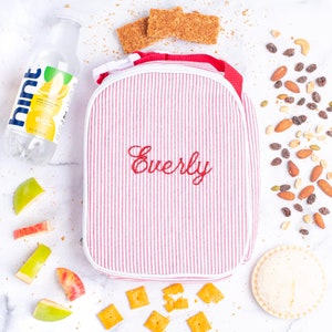 May include: Red and white striped lunch bag with a white trim and the name "Everly" embroidered in red. The bag is sitting on a white surface with various snacks and a bottle of water.