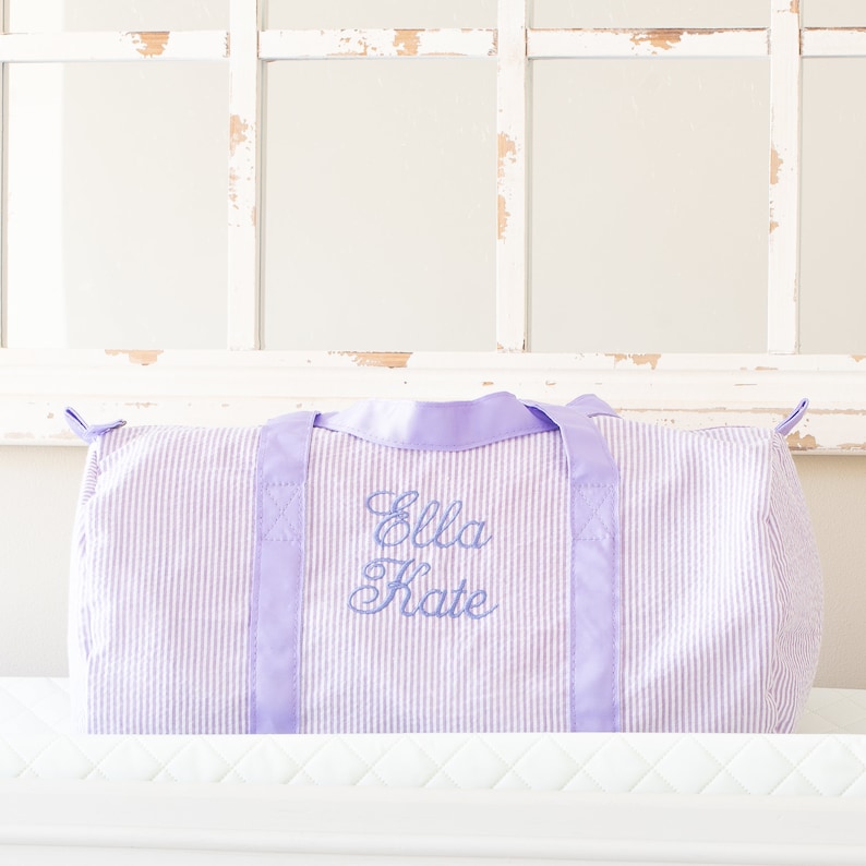 May include: A lavender and white striped duffel bag with a lavender satin handle and the name "Ella Kate" embroidered on the front.