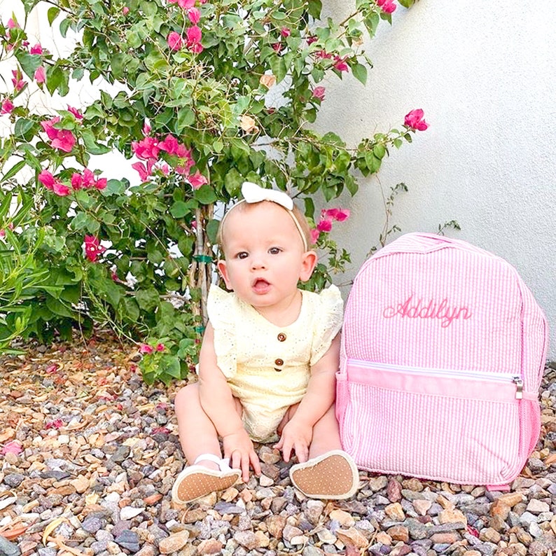 May include: A pink and white striped backpack with the name "Addilyn" embroidered on the front. A baby girl is sitting in front of the backpack, wearing a yellow dress and a white bow.