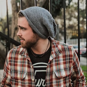 May include: A man wearing a grey beanie, a red and black checked flannel shirt, and a black t-shirt with a white logo. The logo is a circle with the word "Hurley" written in a cursive font.