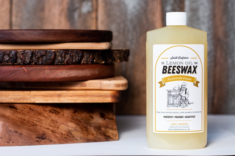 Może przedstawiać: Plastikowa butelka pasty do mebli z wosku pszczelego z olejkiem cytrynowym Amish Craftsmen z białą nakrętką i etykietą z ilustracją stolarza. Tekst na etykiecie to Lemon Oil Beeswax Furniture Polish, Protects, Polishes, Beautifies, 16 fl oz.