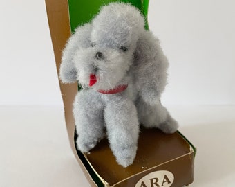 Può includere: Un giocattolo di peluche di barboncino grigio in posizione seduta con un collare rosso e una lingua rossa che sporge. Il giocattolo è confezionato in una scatola marrone con la parte superiore verde e la parola "ARA" stampata sul davanti.