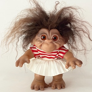 May include: A vintage troll doll with brown hair and large orange eyes. The doll wears a red and white striped top and a white pleated skirt. The troll has a smiling expression and is standing.