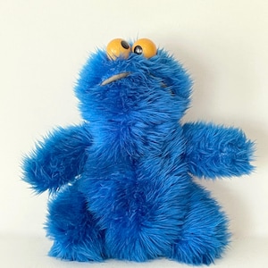 Könnte beinhalten: Ein blaues Plüschtier eines Monsters mit großen gelben Augen und einem breiten Mund. Das Spielzeug hat zotteliges blaues Fell und ausgestreckte Arme, das einem beliebten Kindercharakter ähnelt.