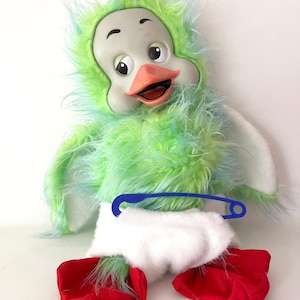 May include: A plush duck doll with a light green and blue shaggy faux fur body, a smooth gray face, and an orange beak. The doll wears a white fabric diaper secured with a large blue plastic safety pin and has bright red fabric feet.