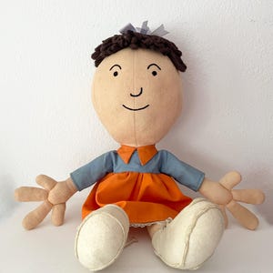 Puede incluir: Una muñeca hecha a mano con una cara de color canela claro, cabello de hilo marrón y un lazo gris. La muñeca viste una camisa azul con un vestido naranja y zapatos blancos. Los brazos de la muñeca están extendidos y tiene una cara sonriente.