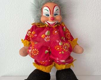 May include: A vintage clown doll with a smiling face, grey hair, and a red floral jumpsuit with yellow trim. The doll has black shoes and tan hands. The clown's face is painted with red, white, and blue accents.