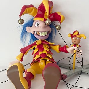 Puede incluir: Un títere de bufón con cabello azul, que lleva un sombrero puntiagudo rojo y amarillo con cascabeles metálicos y una túnica a capas a juego. El títere presenta dientes grandes, una sonrisa amplia y sostiene un bufón más pequeño en una varilla.