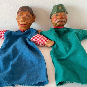 Puede incluir: Dos marionetas de mano de madera con caras pintadas y ropa de tela. Una marioneta lleva una camisa azul y mangas a cuadros rojas y blancas. La otra marioneta lleva una camisa verde y un sombrero verde con ribete dorado.