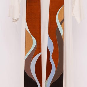 May include: A brown and tan liturgical stole with an abstract design of flowing shapes in shades of brown, blue, and tan. The stole is displayed on a white garment, likely a robe, against a white background.