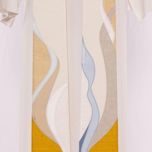 May include: Cream-colored liturgical stole with abstract, flowing designs in shades of gold, beige, white, gray, and blue. The stole has a bright yellow border at the bottom. The stole is worn over a white garment.