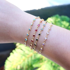 May include: Gold-toned bracelets with heart-shaped gemstones in various colors, including blue, red, black, and pink. The bracelets are linked together with small gold chains. The image shows the bracelets on a wrist.