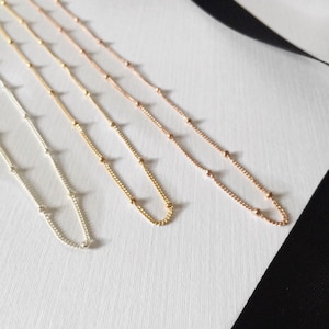 May include: Three delicate gold chains with small beads, one silver chain with small beads, and one rose gold chain with small beads. The chains are laid out on a white surface with a black ribbon.