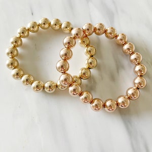 May include: Two gold-toned beaded bracelets, one slightly larger than the other, are intertwined on a white marble surface.