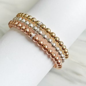 以下が含まれることがあります： ゴールド、シルバー、ローズゴールドのビーズでできた3層のブレスレット。真ん中のブレスレットには、小さなクリアクリスタルアクセントが付いています。